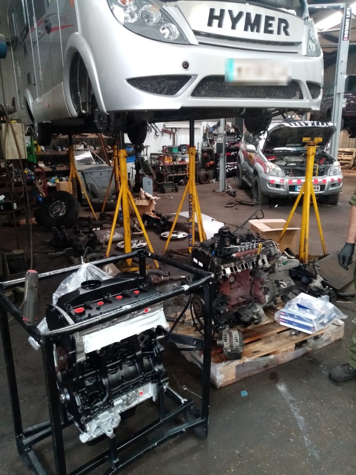 Inside the workshop: a motorhome raised on yellow mobile column lifts, an engine on a stand in front